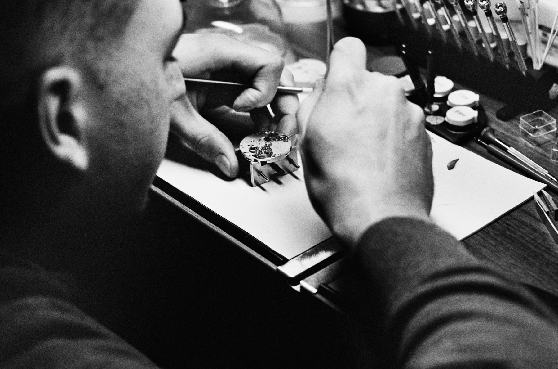 Black and white image of a watchmaker using precision tools to assemble a mechanical watch movement at a workbench surrounded by specialized instruments.