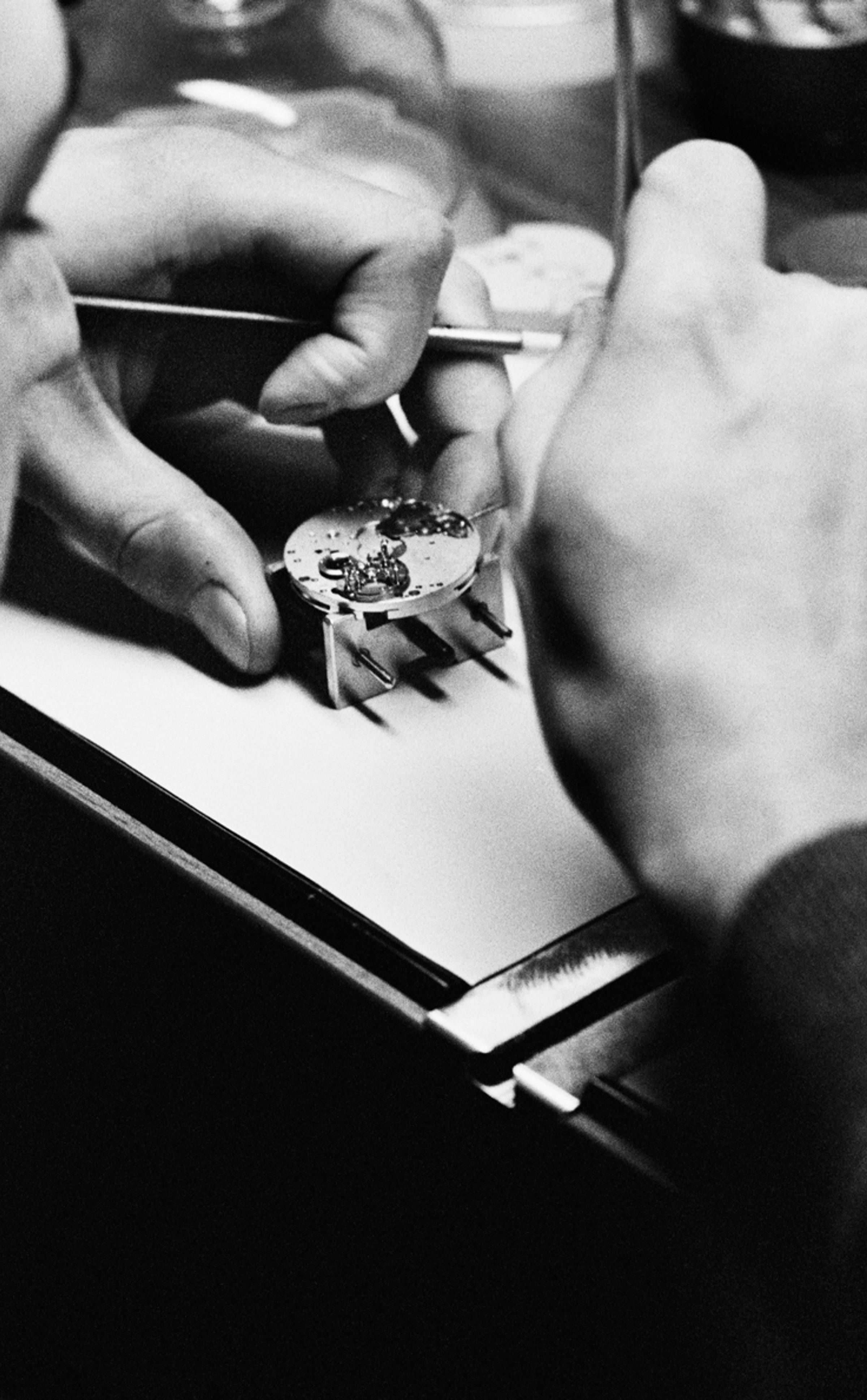 Black and white image of a watchmaker using precision tools to assemble a mechanical watch movement at a workbench surrounded by specialized instruments.