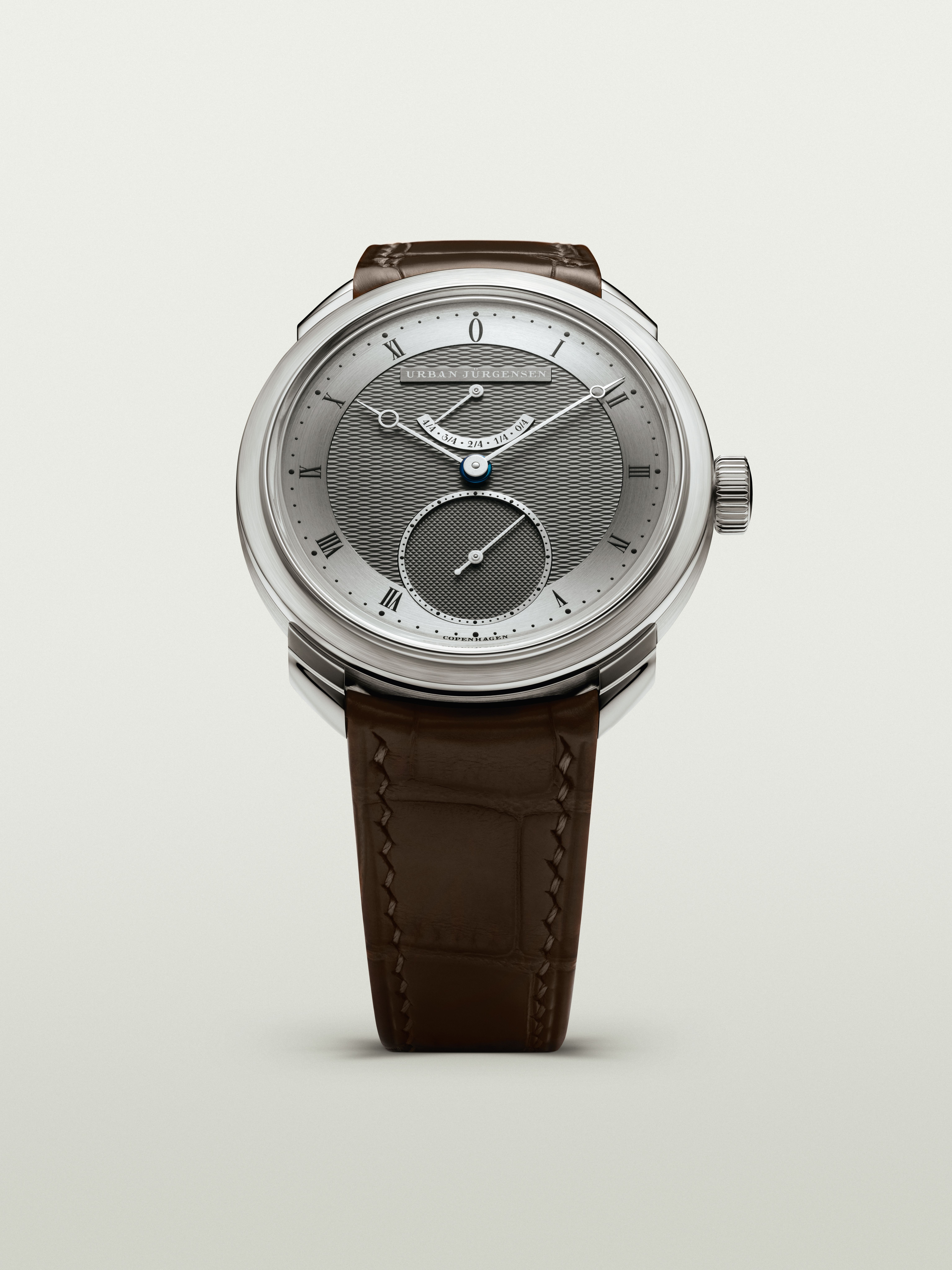 Front view of a platinum Urban Jürgensen watch with dark guilloché dial, blued hands, power reserve indicator, and brown leather strap.