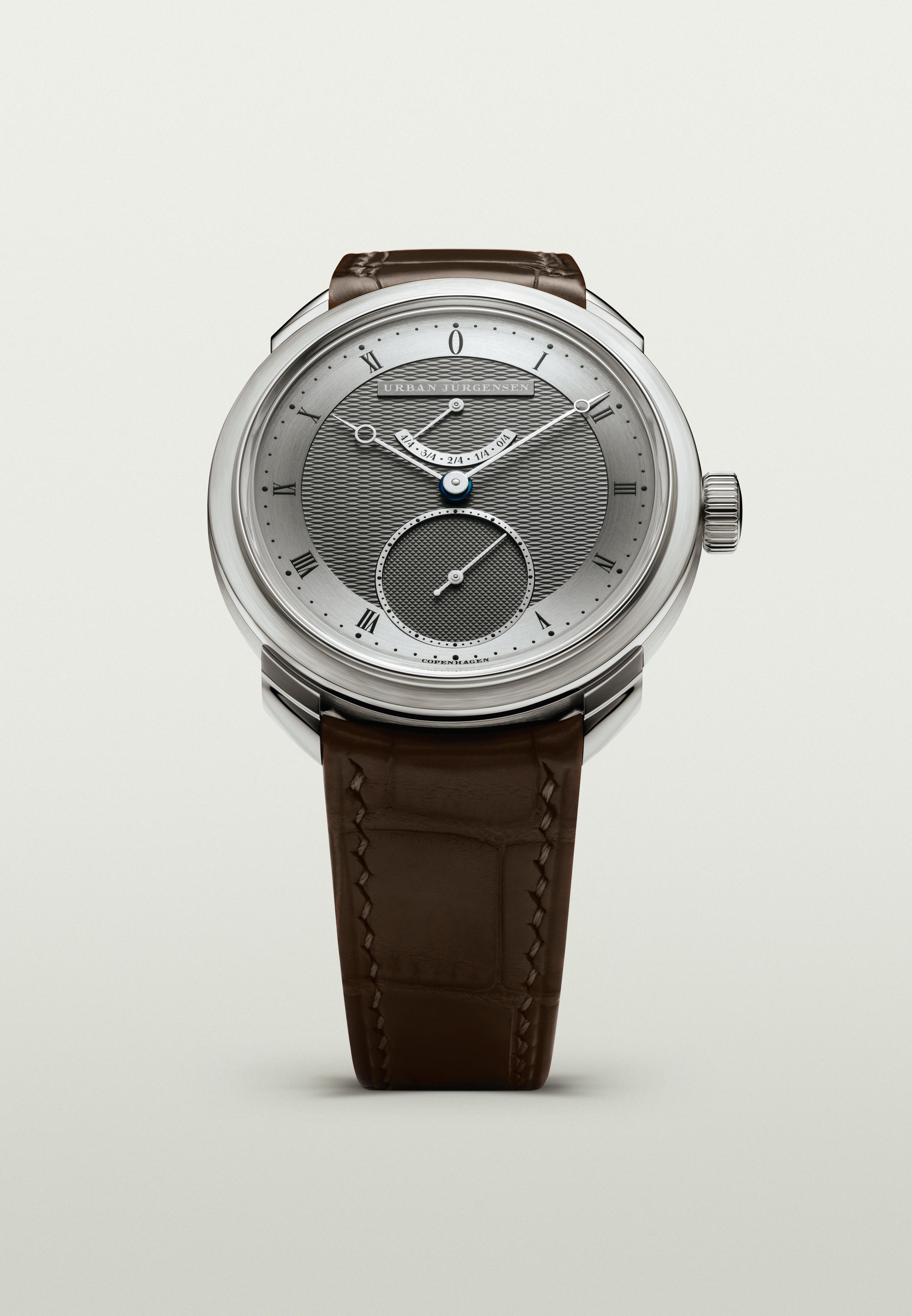 Front view of a platinum Urban Jürgensen watch with dark guilloché dial, blued hands, power reserve indicator, and brown leather strap.