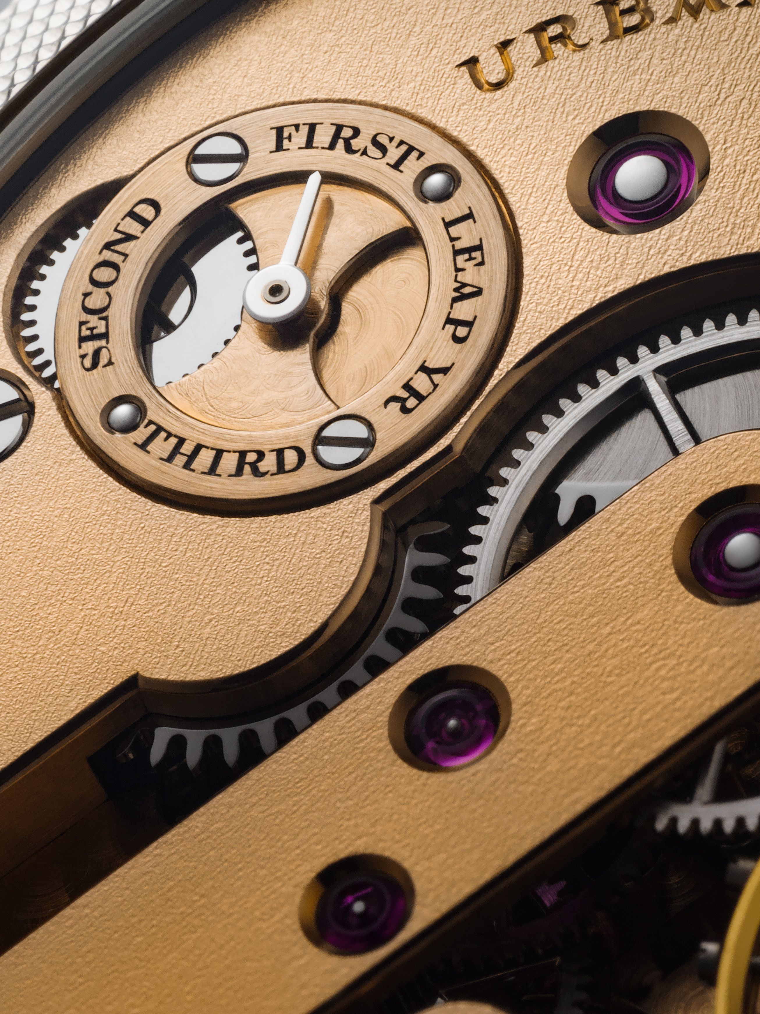 Close-up of a rose gold leap year indicator on a watch movement, showing engraved labels for First, Second, Third, and Leap Yr, with visible gears and jewel bearing.