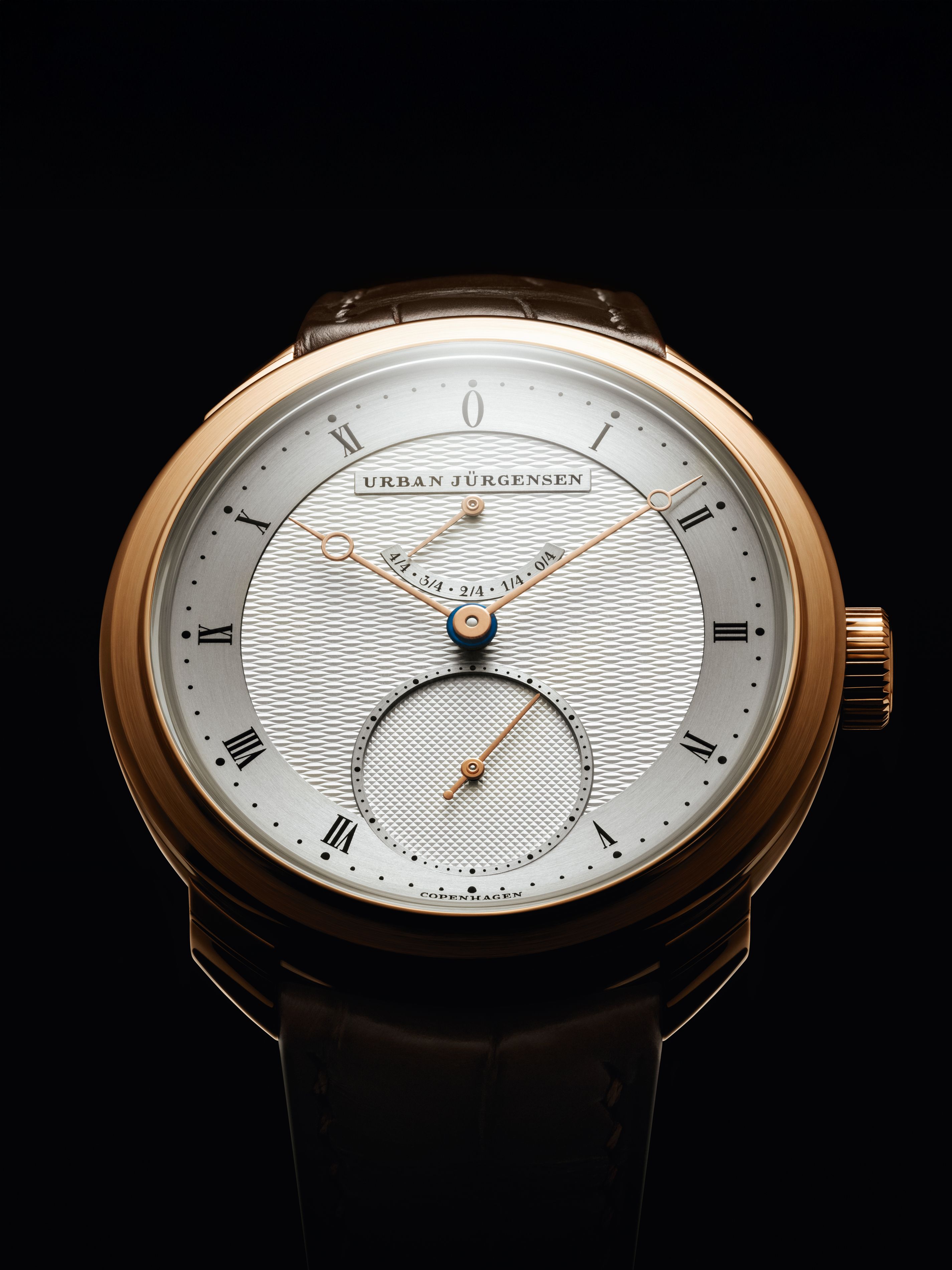 Front view of a rose gold Urban Jürgensen watch with silver guilloché dial, Roman numerals, and small seconds subdial, set against a black background.