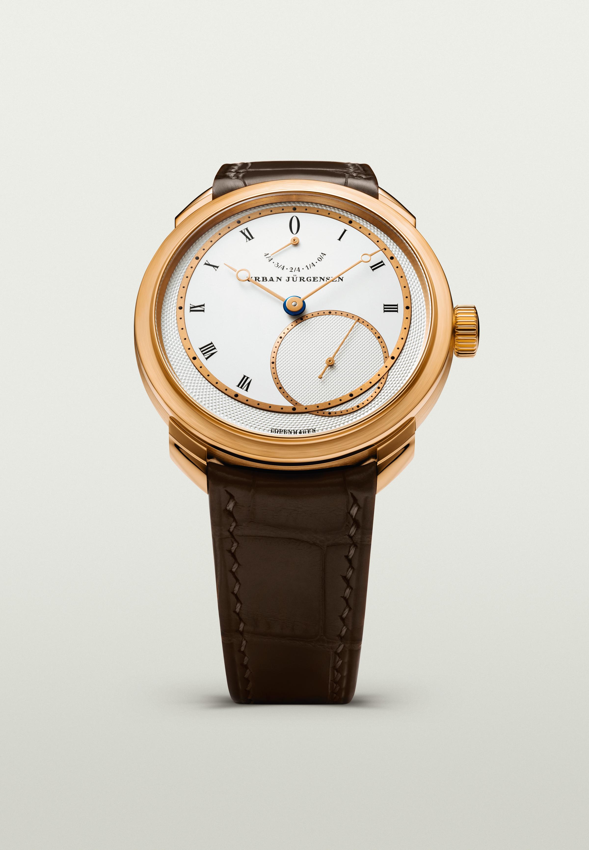 Rose gold Urban Jürgensen wristwatch with silver dial, gold hands, Roman numerals, power reserve indicator, and brown leather strap.