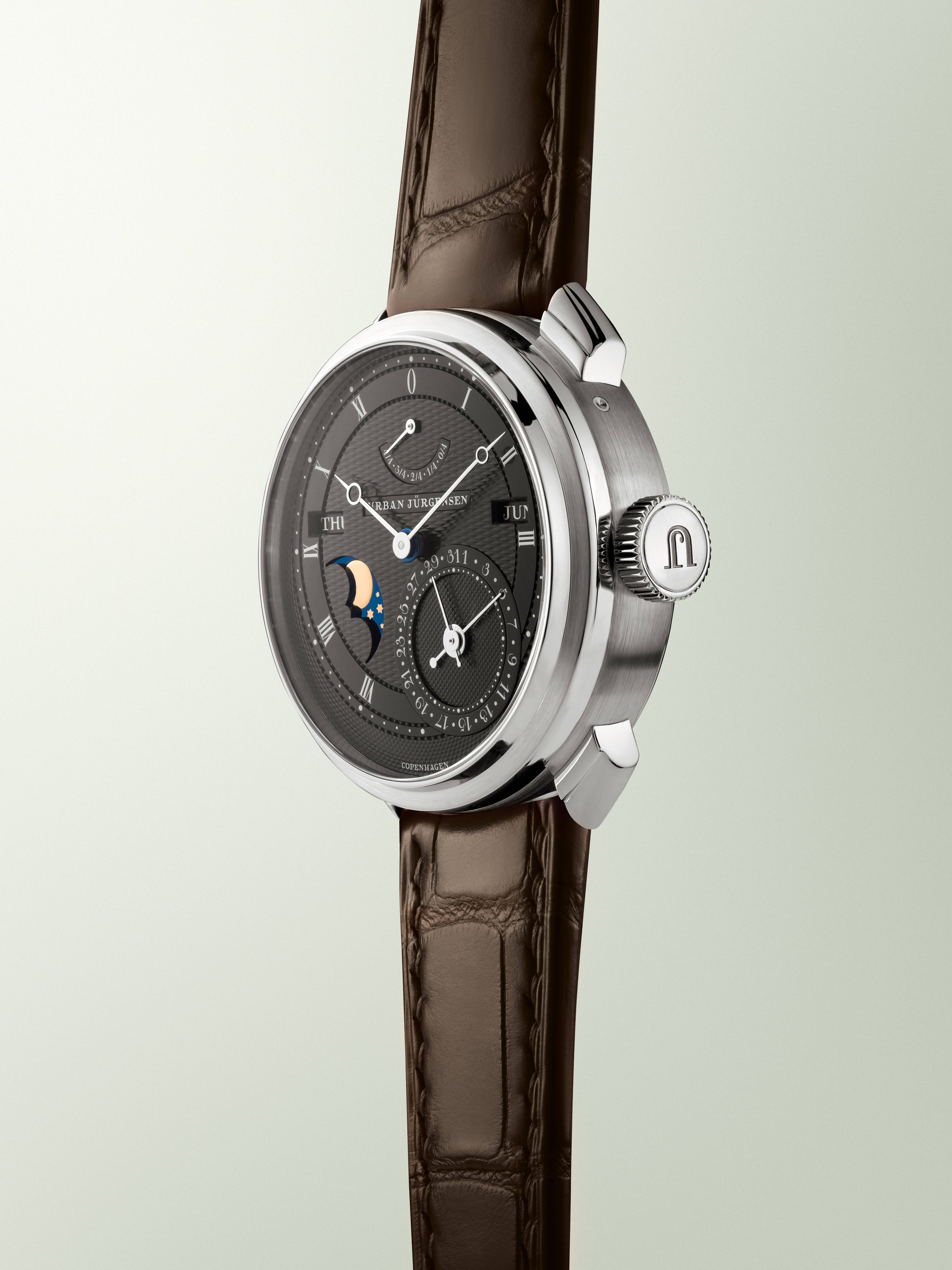 Platinum Urban Jürgensen wristwatch with dark guilloché dial, moonphase, day and date indicators, power reserve display, and brown leather strap.