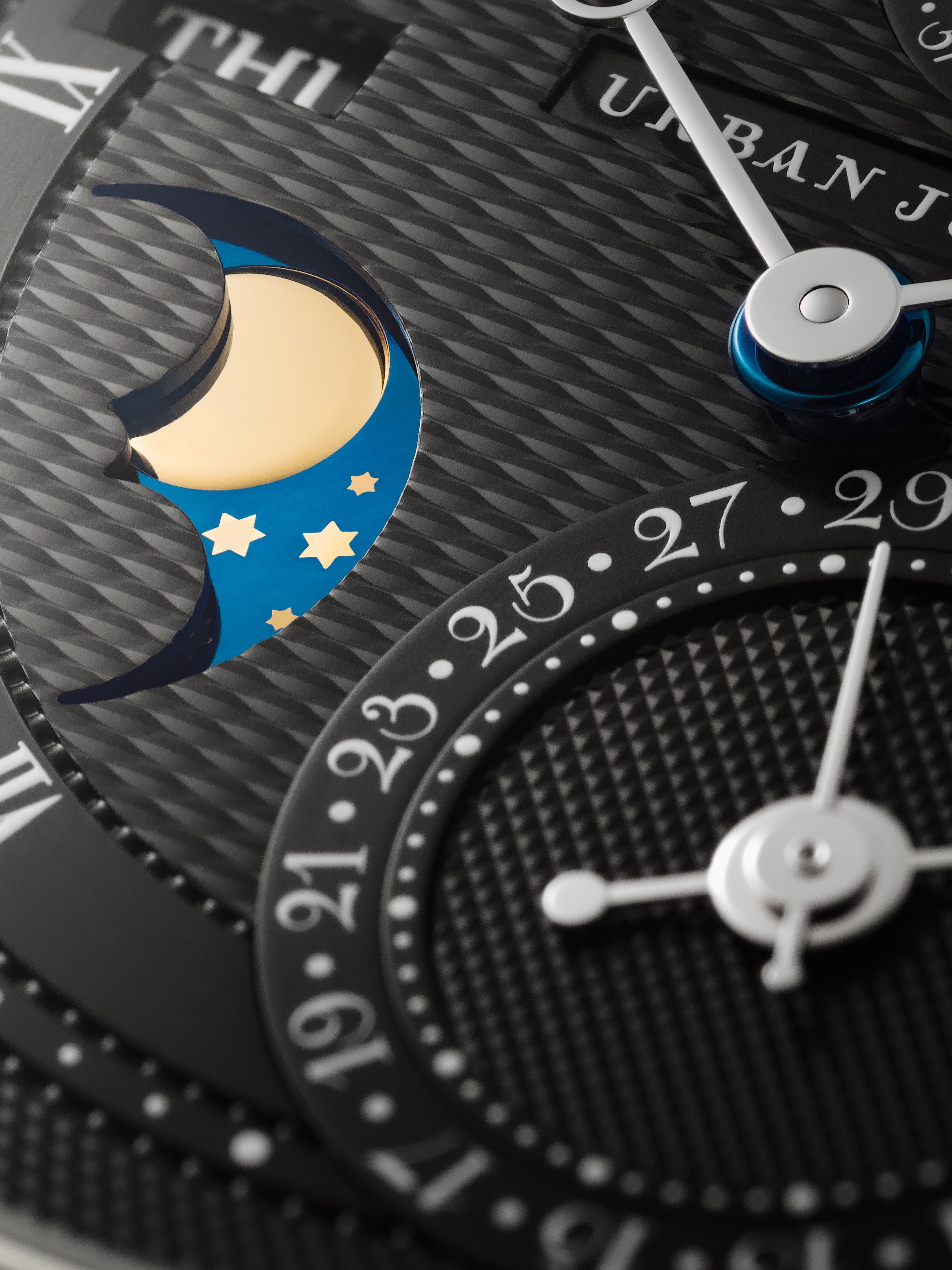Close-up of a black guilloché watch dial featuring a moonphase display with gold moon and stars on blue, date subdial, day window, and polished hands.
