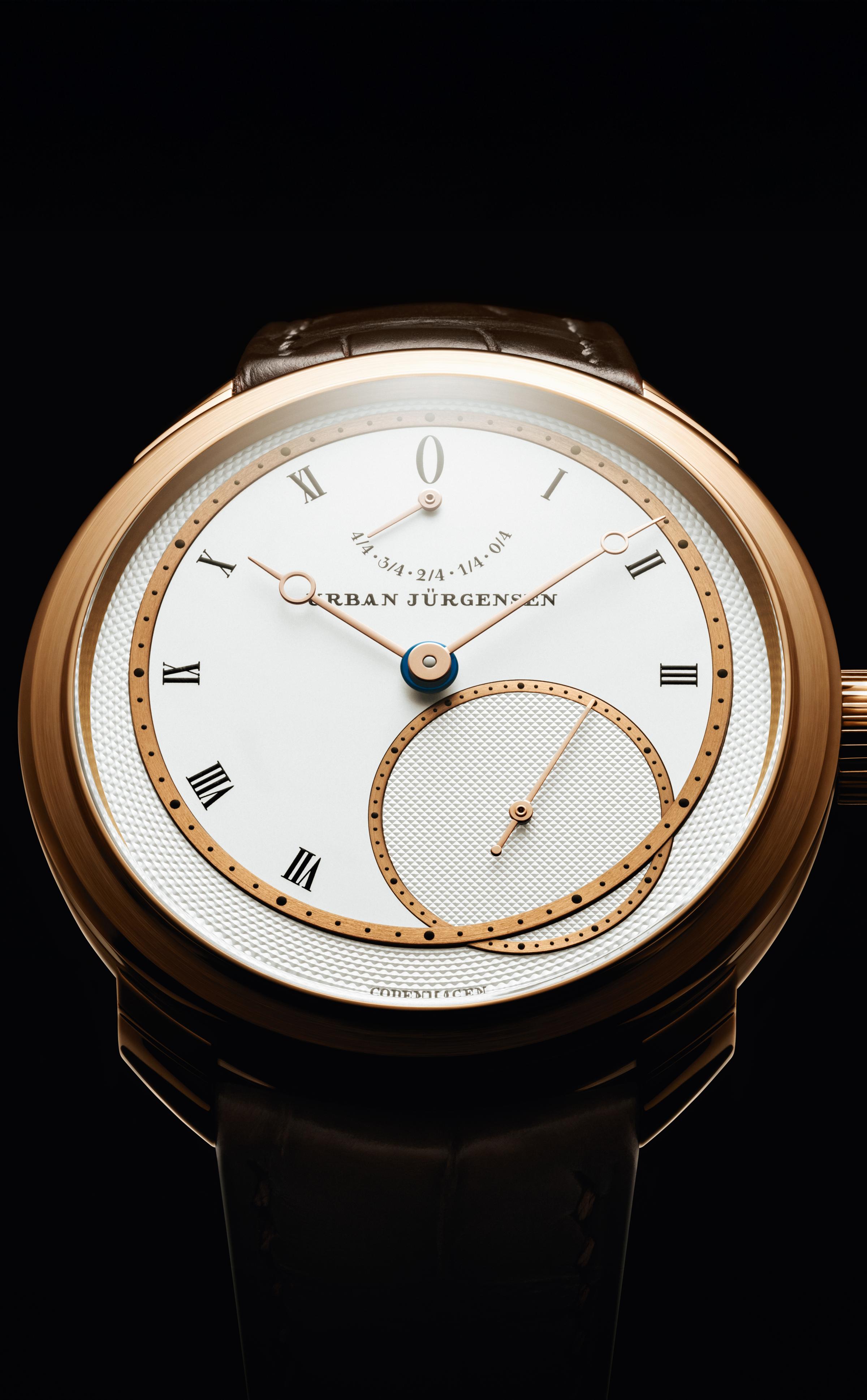 Front view of a rose gold Urban Jürgensen wristwatch with silver dial, gold hands, Roman numerals, power reserve indicator, and small seconds subdial, set against a black background.