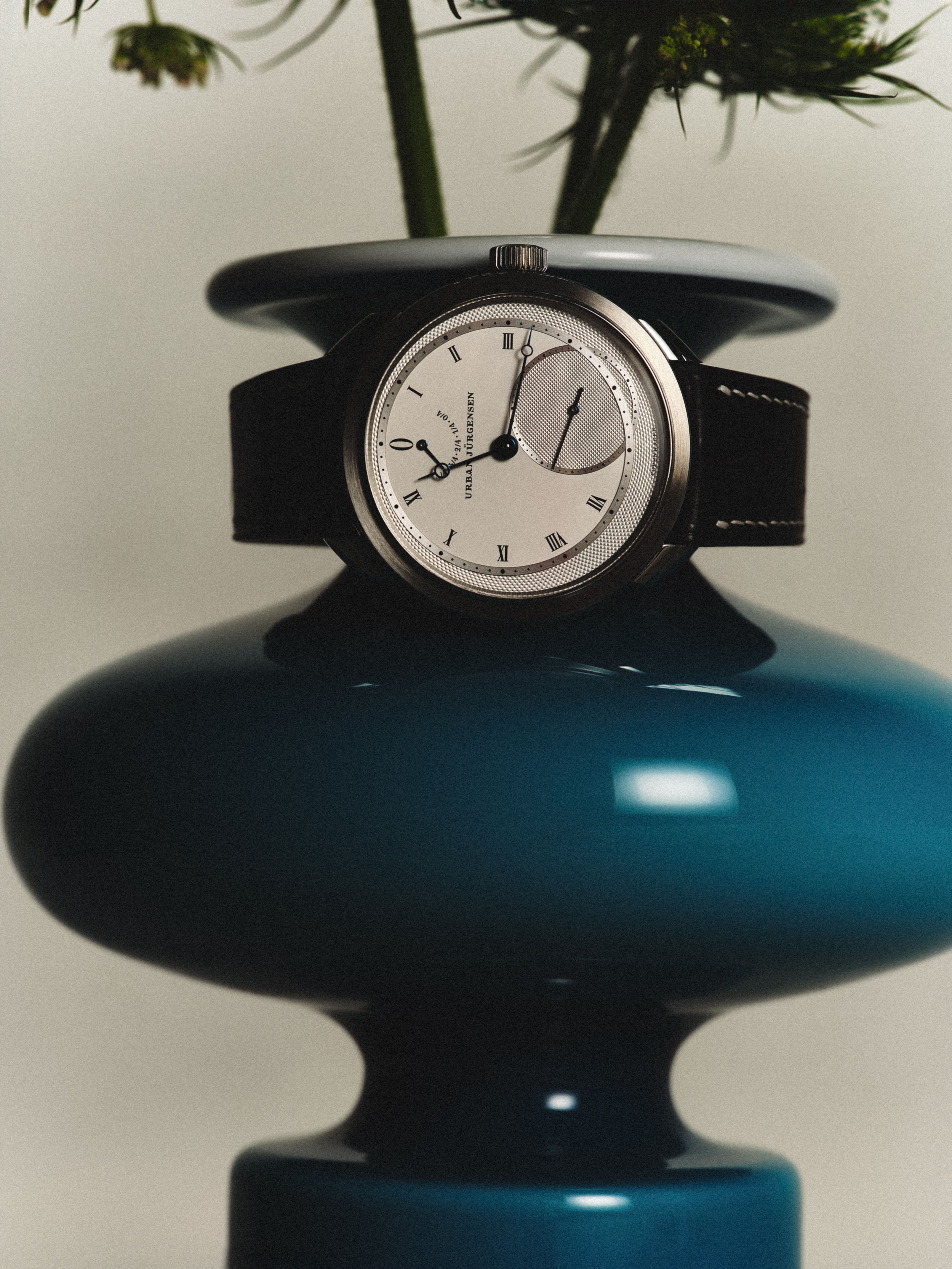 Urban Jürgensen wristwatch with silver dial and black strap displayed on a glossy dark teal vase, with green flower stems emerging from the top.