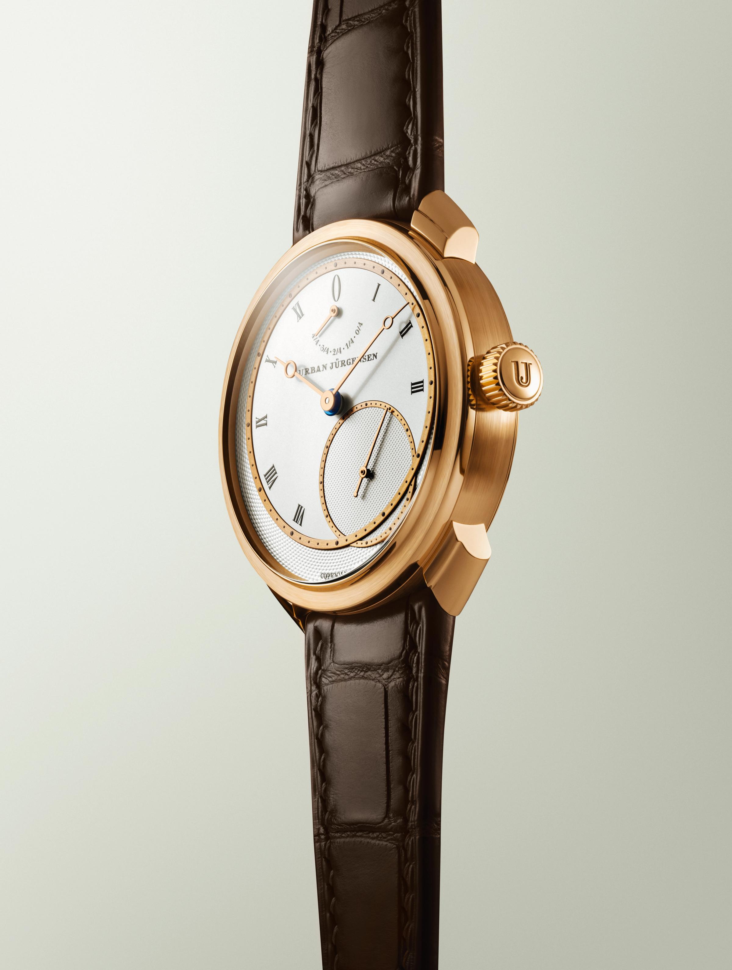 Rose gold Urban Jürgensen wristwatch with silver dial, gold hands, Roman numerals, power reserve indicator, and brown leather strap.
