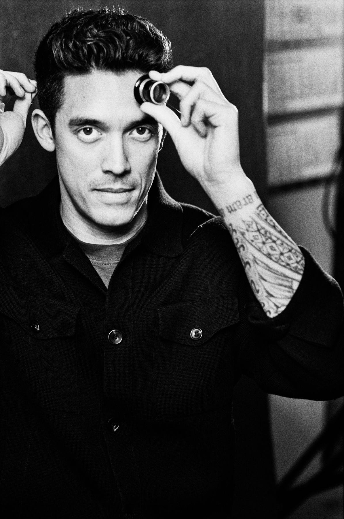 Portrait of a watchmaker in a dark shirt, holding a loupe over one eye and looking directly at the camera, with a tattooed forearm partially visible.