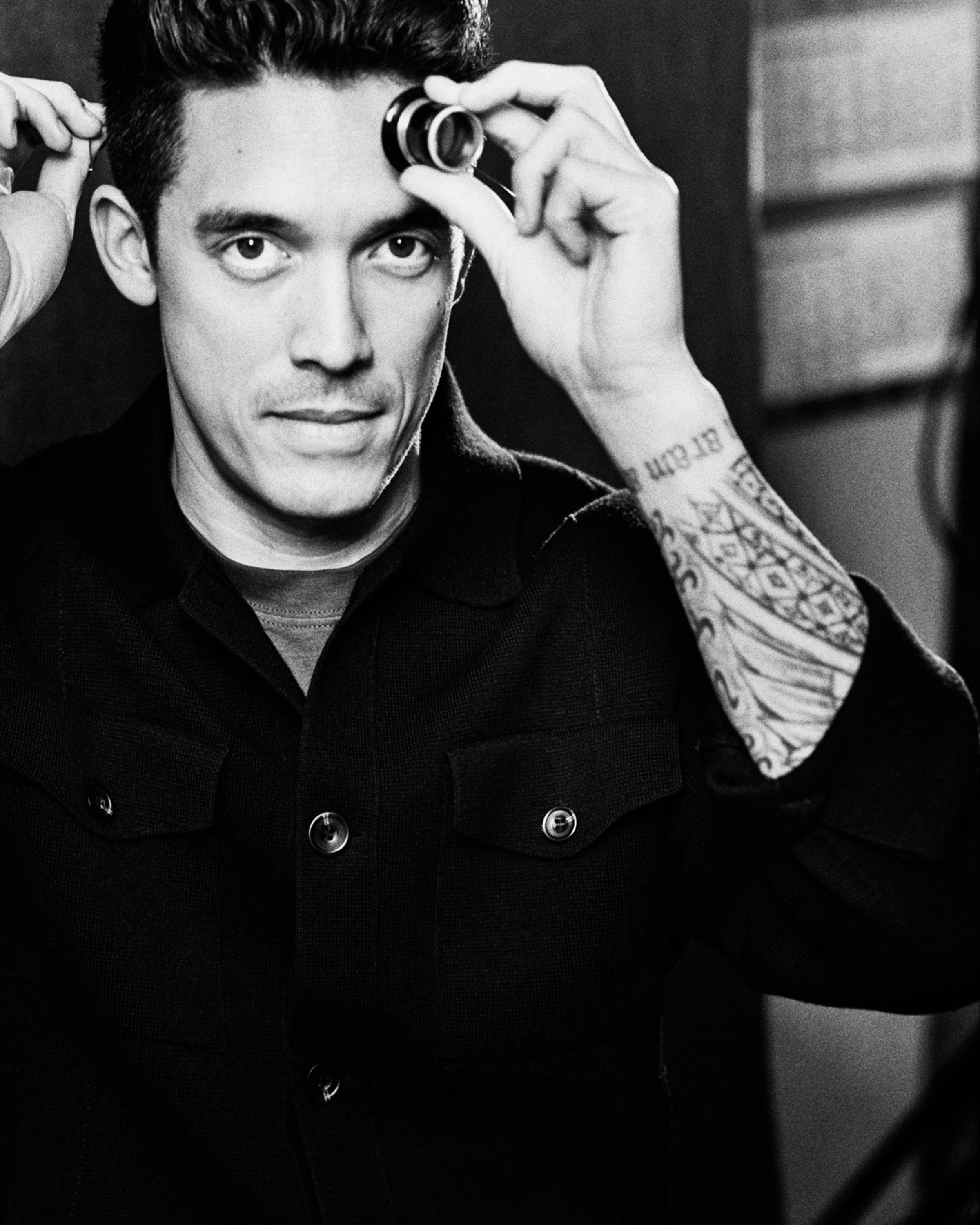 Portrait of a watchmaker in a dark shirt, holding a loupe over one eye and looking directly at the camera, with a tattooed forearm partially visible.